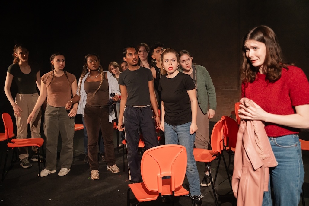 Action shot of the spring 2024 Shakespeare in Performance at RADA final presentation of Julius Caesar. An actor wearing a bright red shirt stands on the right, holding a light pink jacket, with a concerned expression on her face. A group of actors stands to the left with concerned expressions on their faces.